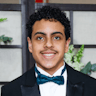 A boy with curly hair smiling, wearing business clothes with a blue bow tie.